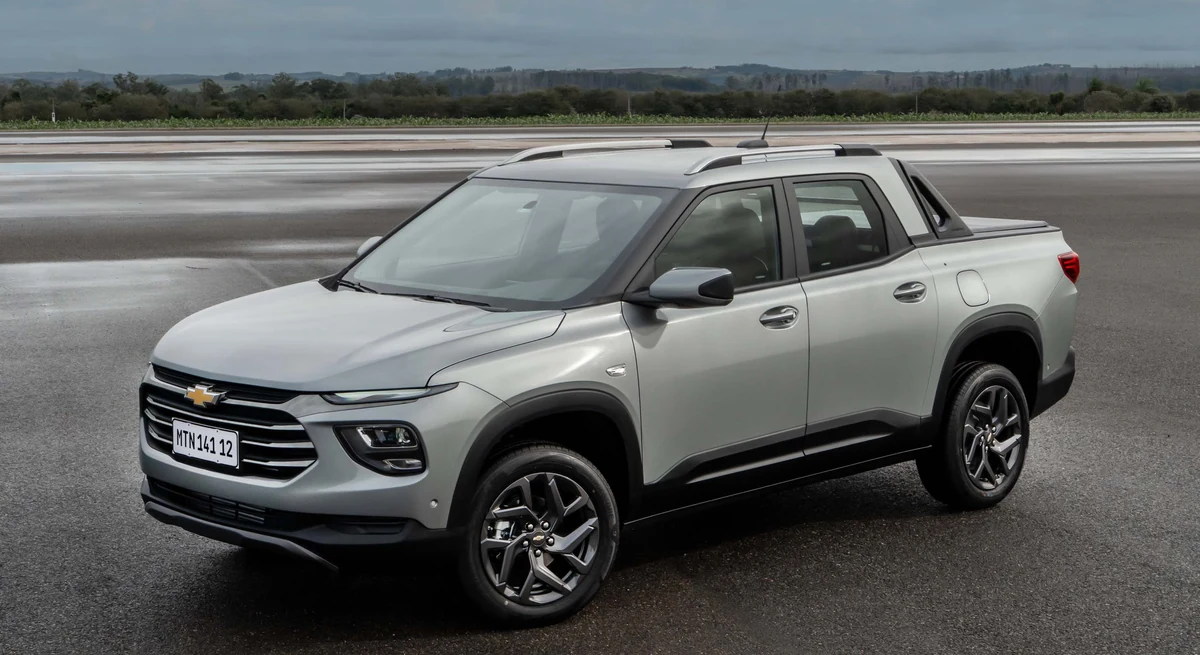 Chevrolet Montana Premier da linha 2027, na cor prata, fotografada em três quartos dianteiro sobre pista de asfalto molhado, com vegetação e céu nublado ao fundo