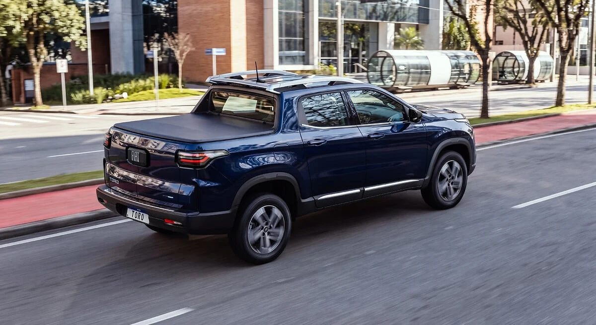 Fiat Toro 2027 azul em movimento numa avenida urbana, vista traseira em três quartos