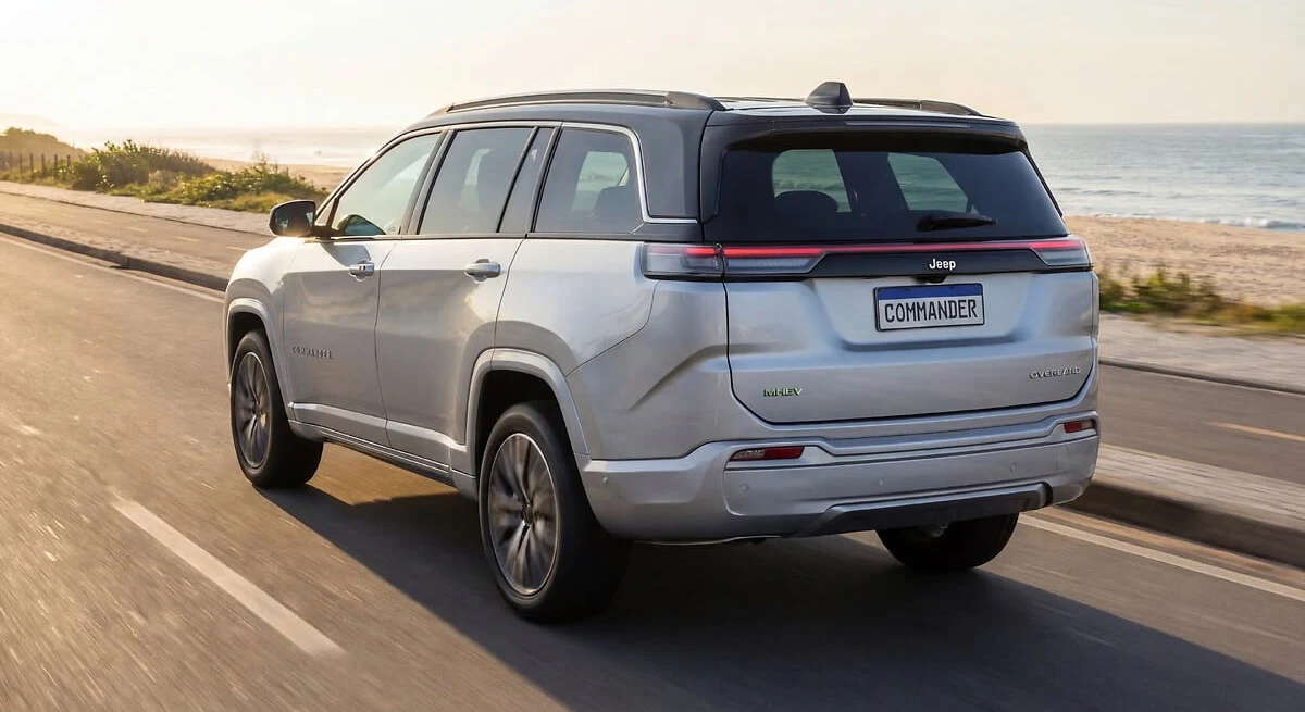 Jeep Commander 2027 prata em movimento numa estrada à beira-mar, vista traseira em três quartos com o emblema MHEV na tampa traseira