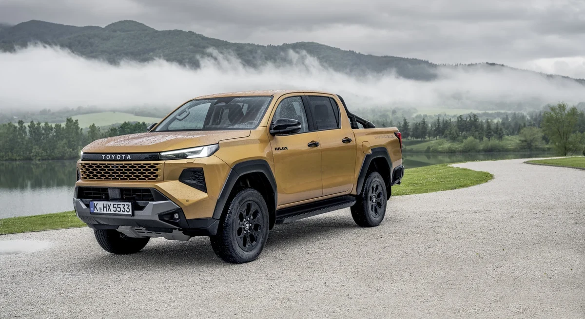 Nova Toyota Hilux Hybrid 48V amarelo mostarda parada em três quartos dianteiro numa estrada de cascalho, com montanhas e neblina ao fundo